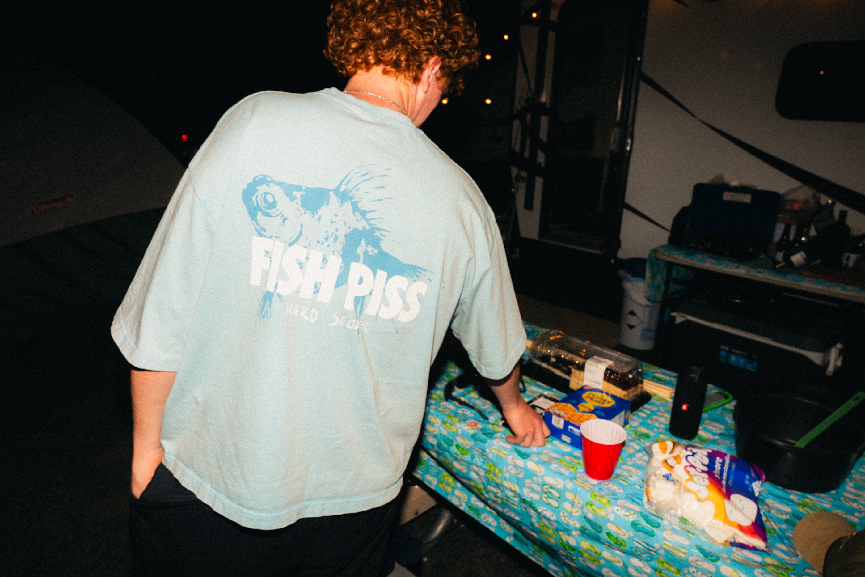Person wearing a light blue t-shirt with text, standing near a table with snacks and drinks at night.